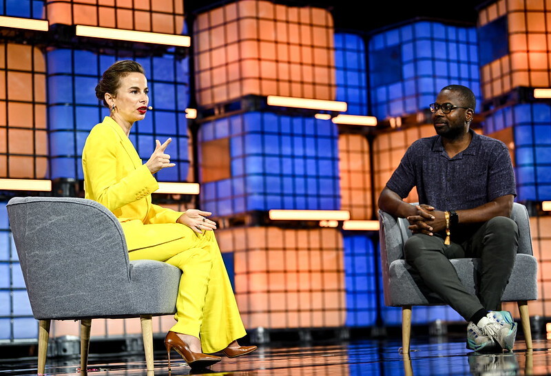 woman in bright yellow suite sitting and talking on a stage at Web Summit Vancouver in front of a blue and orange background