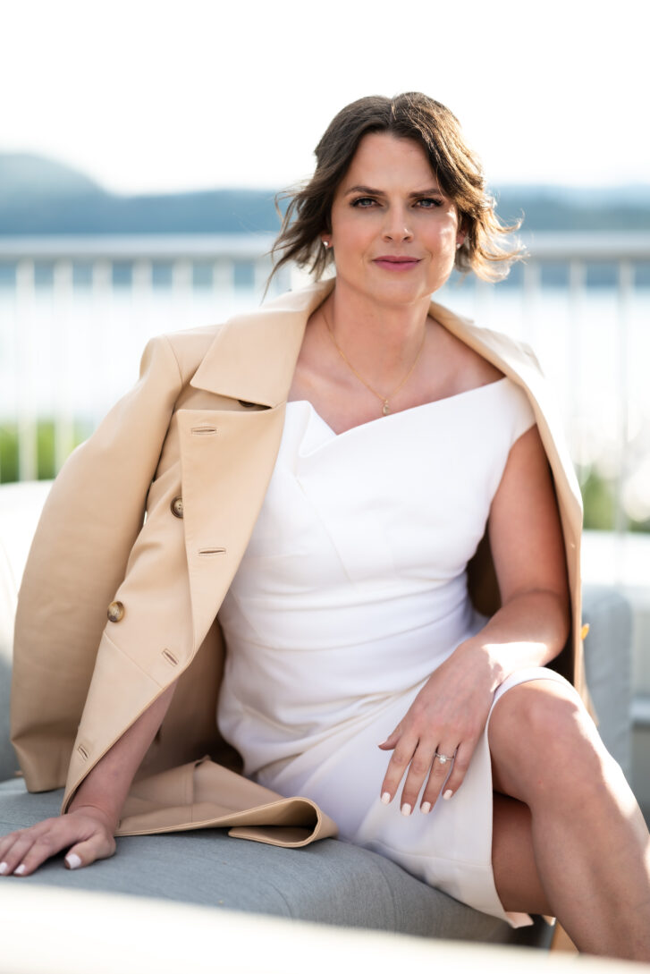 Reece Tomlinson wearing a beige business jacket and white dress, a lake and mountains are behind her in the distance.